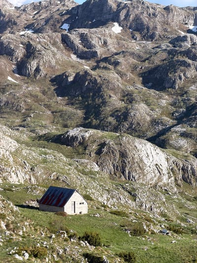 A mountain scene with a small building. The building is perched on a steep, grassy slope, with rugged, rocky mountains on either side. The building has a grey stone structure with a dark red or blue roof. The area around the building is covered in green grass and small shrubs, contrasting with the bare rock. The mountains are rugged and rocky, with patches of green moss and some snow. The image gives a sense of remoteness and harsh environment. Location is Boričje, Montenegro.