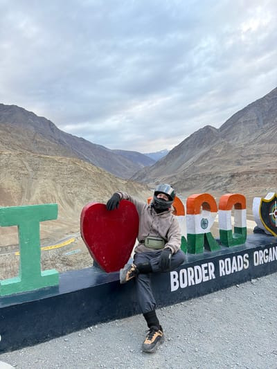 A person in a motorcycle helmet and casual clothing sits on a large, colorful sign in Chumkar, India, with a mountainous landscape in the background. The sign, which appears to spell out "I LOVE INDIA" with a heart in place of "LOVE," is adorned with the Indian flag colors and text that reads "BORDER ROADS ORGAN." The person is wearing a gray hoodie, black pants, black gloves, and hiking shoes. A green fanny pack is worn across their chest. The person's right leg is extended, and their left leg is bent, with their left foot resting on the sign. The environment is characterized by dry, rugged mountains and a cloudy sky. The ground in front of the sign is covered in gravel.