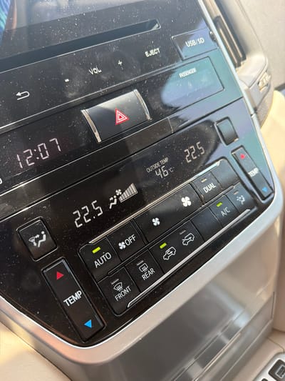 The image provides a close-up, slightly tilted view of a car's dusty center console, highlighting the climate control and infotainment system. No people are visible in the frame.
The digital display prominently shows an extremely high outside temperature of 46°C, indicating very hot weather conditions consistent with the Umm Salal Muhammad, Qatar location. The set interior temperature is 22.5°C, and the current time is 12:07.
The climate control panel indicates several active functions: the A/C is on, and the system is in AUTO mode, both confirmed by green indicator lights. The fan speed is shown to be high, with multiple bars illuminated. Buttons for "DUAL" zone control, "OFF," and "TEMP" adjustment (with red and blue arrows) are visible. Both the "FRONT" and "REAR" defogger/heater buttons are also illuminated green.
On the infotainment section, labels such as "USB/SD," "PASSENGER," and "EJECT" are visible, along with "VOL" for volume control (marked with '+' and '-'). A large red triangular button for hazard lights is centrally located. The overall surface of the console appears to have a fine layer of dust.