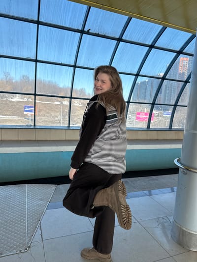 This image depicts a young female posing inside an elevated public walkway or structure, identified as being located in Nikol'skii, Russia.
**Scene Overview:** The indoor space features a tiled floor, a patterned metal ramp section, and a distinctive curved glass wall forming a large window grid. The exterior view reveals a clear blue sky, a winter or early spring landscape with bare trees and snow patches, and a modern urban environment with tall buildings and roads.
**Subject:** A light-skinned female with light brown hair is centered in the frame, facing slightly away from the camera and looking back over her right shoulder with a smile. She is wearing a black long-sleeved top, a black and white houndstooth pattern puffer vest, black wide-leg pants, and light brown, lug-soled boots. Her right leg is lifted and bent, with the sole of her boot prominently displayed.
**Indoor Environment:** The floor is tiled in a light grey pattern, with a silver diamond-plate metal section on the lower left. A low barrier, painted blue on its lower portion and topped with lighter tiles, runs along the base of the windows. A grey cylindrical pillar with a silver handrail is present on the right side. The ceiling above the windows is a light yellow or beige color. The large, multi-paneled glass windows are slightly streaked with what appear to be water marks or grime.
**Outdoor Environment (visible through windows):**
* **Left Side:** A sloping, snow-dusted or dry grassy hillside with numerous bare deciduous trees. A road and a blue billboard with white Russian text, partially visible as "Денег нет..." (No money...), are discernible in the distance.
* **Right Side:** A dense urban landscape featuring multiple tall, modern high-rise buildings, including one with a distinct orange-red facade. A multi-lane road with vehicle traffic is visible, along with a large pink billboard displaying white Russian text that reads "РУКИ ВВЕРХ" (Hands Up!). The sky above is clear and blue.
