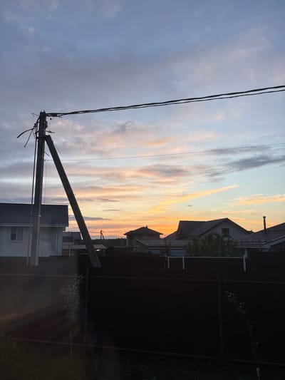 This media file captures a sunset scene in Дольче Вита Кп, Russia. The sky is filled with dramatic clouds painted in hues of orange, pink, and yellow, indicating the time of day is late evening. A prominent power line pole and wires dominate the foreground, stretching across the frame. In the background, residential houses with dark roofs are visible, suggesting a suburban or village setting. A dark fence separates the properties. The lighting suggests the sun is low on the horizon, casting a warm glow on the clouds. The scene evokes a sense of calm and the transition from day to night. No people are visible in the frame, and there is no visible text.