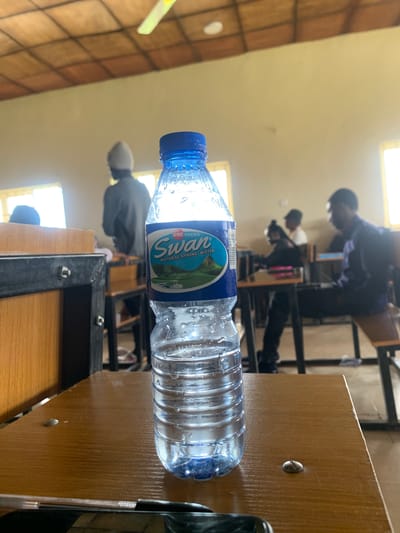 A close-up, low-angle shot captures a partially filled plastic water bottle resting on a wooden surface in what appears to be a classroom or study hall setting. The bottle has a blue cap and a label that reads "Swan Natural Spring Water" from "UAC FOODS". The water inside is clear, with condensation visible on the bottle's exterior.
In the background, out of focus, several individuals are seated at desks, suggesting an educational environment. The lighting indicates it could be daytime, with light streaming in from windows. The overall impression is one of a casual moment during a study session or class. The wooden surfaces of desks and possibly shelving are visible, along with the metallic frames of the desks. The scene is set in Farin Yashe, Nigeria.