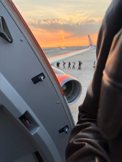 The image depicts an airport tarmac scene at sunset, observed from within an aircraft at Bordeaux-Mérignac Airport (BOD) in Mérignac, France. The perspective is through an open aircraft door or large window, with the light grey interior paneling, including a black latch mechanism and fasteners, visible on the left. A dark brown, textured object, likely a jacket sleeve or gloved arm, partially obstructs the right side of the view.
Immediately outside the aircraft, the white wing with an orange stripe and the front of a jet engine nacelle, showing fan blades, are visible. On the grey asphalt tarmac below, a line of approximately six to seven individuals, dressed in dark clothing, are walking in single file from left to right across the frame. Further to the right, another individual or small group is present. In the mid-distance, the tail section of an aircraft with an orange vertical stabilizer, identifiable as an EasyJet livery, is visible. Other indistinct aircraft and airport infrastructure are present on the distant tarmac.
The sky is illuminated by a sunset, exhibiting strong orange and yellow hues near the horizon, transitioning to lighter tones above. Scattered dark clouds are present. The setting sun is a bright, obscured orb near the horizon line. The overall lighting is warm, indicative of golden hour conditions.
