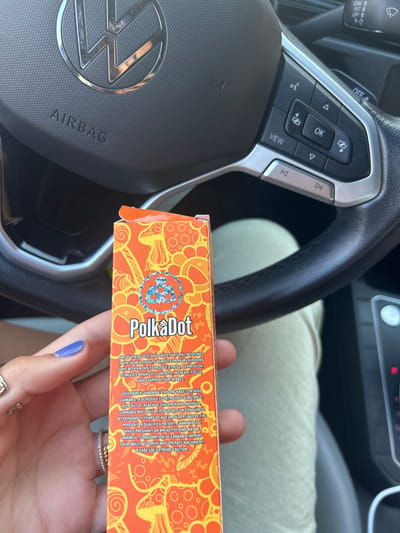 Inside a car, a person holds a "Polka Dot" mushroom chocolate box. The box claims it contains one of the oldest and safest natural medicines. It has health benefits from mushrooms that reduce stress, depression, stimulate brain cell growth, increase focus, and sharpen senses. A warning label indicates the package contains cannabis, which is a schedule-controlled substance. It is for adults 21 years or older unless the person is a qualified patient. It warns about impaired ability to drive and operate machinery.