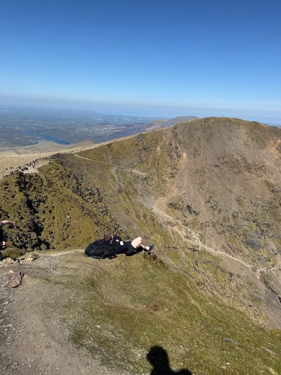 A high-angle, landscape-oriented photograph depicts a mountainous region under a clear, blue sky, likely taken near Beddgelert in the United Kingdom. A person, partially visible in the lower center, is positioned near the edge of a steep slope. They appear to be wearing dark clothing and possibly carrying a backpack.

Further down the mountain, a discernible path is visible, lined with numerous small figures, suggesting a popular hiking trail. In the distance, a body of water is nestled among the rolling hills and flatlands stretching to the horizon. The terrain is a mix of rocky outcroppings, sparse vegetation, and patches of grass. The image exhibits a tilted perspective, likely due to the camera's orientation at the time of capture.