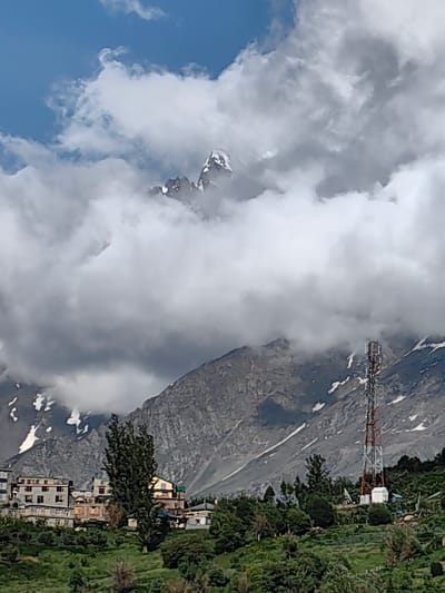 The image depicts a mountainous landscape in Kardang, India, with a village nestled at the foothills. The scene is dominated by a towering, snow-capped peak emerging from a dramatic bank of clouds, suggesting a high-altitude location. The weather appears to be partly cloudy, with the sun intermittently breaking through, casting a soft light on the terrain.

In the foreground, a cluster of buildings forming the village are visible. These structures are low-rise, with multiple windows and some appearing to be residential or guesthouses. A tall, red and white telecommunications tower stands prominently on the right side of the image, indicating modern infrastructure within the remote setting. Lush green vegetation, including trees and possibly agricultural fields, covers the lower slopes, contrasting with the rocky, barren mountainside above. Snow patches are visible on the upper slopes, reinforcing the mountainous environment.

The overall atmosphere is one of natural grandeur and a sense of human settlement integrated into a rugged landscape. There is no discernible activity or event taking place, and no visible text in the image. The time of day appears to be daytime, possibly late morning or early afternoon, given the lighting conditions.