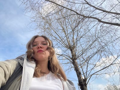 A woman with long blonde hair and glasses is looking down at the camera. She's wearing a white shirt and a grey jacket. Behind her is a tree with bare branches against a light blue sky with some white clouds. The location appears to be outdoors during the daytime. There is no evidence to suggest that the image was taken in Fabriki 1 maya, Russia.