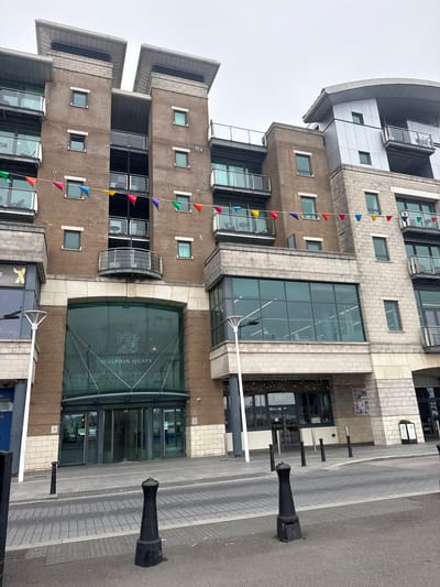 This is a vertical, eye-level photograph of a modern building with a series of windows and balconies. The building's facade is a mix of light-colored brick and large glass sections. A string of colorful triangular flags hangs down the side of the building. The flags are red, yellow, purple, and green, and they are spaced out along a string.
In front of the building, there is a paved walkway and a road. Several black, bollard-like objects are placed along the edge of the walkway. The sky is overcast and appears to be a light grey color, suggesting a cloudy day. The building has multiple levels, with different architectural styles visible. The top section of the building features a curved, silver metallic roof.
One prominent glass entrance has "DOLPHIN QUAYS" written in white lettering, along with a graphic of a fish. There is also a sign with a yellow "X" logo visible below the main entrance. The overall scene suggests an urban environment, possibly a commercial or residential complex. The lighting is diffused, characteristic of an overcast day. No people are clearly visible in the frame. The time of day is not definitively indicated but appears to be daytime.