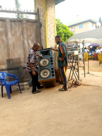 Two men are lifting a large, worn-out speaker cabinet with two large speakers. One man wears a patterned shirt and dark pants, the other a track suit. Behind them is a metal gate, and behind the second man is a sound system setup with equipment on stands. In the background, people wearing yellow skirts and white blouses stand under tents. A blue plastic chair sits to the left of the speaker. The ground is dirt or sand.
