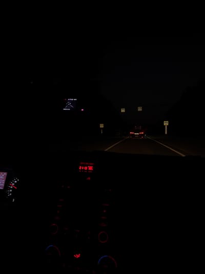 The image is taken from inside a vehicle at night, looking out onto a dark, unlit highway in Letnevo, Russia. The dashboard of the car is visible in the foreground, with a speedometer showing a speed of approximately 60 km/h and a digital display showing the time as 21:13 and the outside temperature as 22°C. The road ahead is dark, with the headlights of other vehicles visible in the distance. There are several yellow road signs with black symbols, indicating potential hazards or directions. The environment appears to be rural or suburban, with no visible buildings or infrastructure other than the road and signs. The overall mood of the image is one of solitude and quiet, with the darkness of the night and the faint glow of the car's instruments creating a sense of introspection.