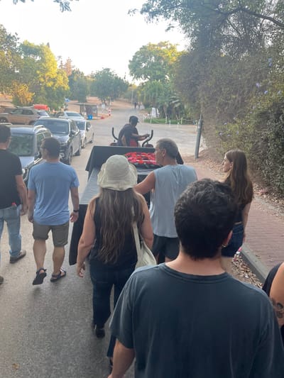 The image portrays an outdoor procession occurring on a paved road within the Brener Regional Council, Israel. A dark-colored utility vehicle, driven by a man wearing a dark t-shirt and cap, moves forward in the midground. The vehicle's rear platform supports a black-draped object topped with a rectangular arrangement of red flowers. A group of six individuals follows directly behind the vehicle, walking on the paved surface. From the foreground to the midground, these individuals include: a man with dark hair in a dark grey t-shirt (foremost), a woman with long brown hair wearing a black top and denim shorts (right side), a man in a light blue tank top and dark shorts (right side), a woman with long brown hair wearing a wide-brimmed light hat, black tank top, and blue jeans (center), a man in a light blue t-shirt and olive shorts (left side), and a man in a dark t-shirt and blue jeans (far left).
On the left side of the road, a line of parked passenger vehicles is visible, including a silver SUV, a dark sedan, and a white compact car. The environment is characterized by dense green foliage, with trees and shrubs lining both sides of the road. A thick hedge with small blue flowers borders the right side of the path. In the distant background, an unpaved road ascends gently, with further trees and a visible archery target and signs on the left. The scene is illuminated by natural daylight, casting soft shadows.