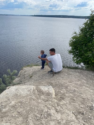 An eye-level shot captures a man and a young boy on a rugged cliff edge, looking out over a vast body of water under a partially cloudy sky. The man, wearing a white t-shirt and grey shorts, is crouched down on the rocky ground, his head turned slightly towards the boy. The boy, dressed in a blue long-sleeved top and dark trousers, stands next to him, also looking downwards, presumably at the water or what's below the cliff.

The cliff edge is composed of light grey, rough earth and scattered rocks and pebbles. A large green tree with leafy branches overhangs from the top left, partially framing the scene and casting some shade. Below the cliff, a wide expanse of water, likely a river or large lake, stretches across the frame, showing ripples on its surface. In the distance, on the far bank of the water, a thin strip of land is visible, featuring a line of trees and what appears to be a distant settlement or industrial structures. The sky above the distant horizon is bright with scattered white clouds.

The scene suggests a daytime setting, possibly late morning or early afternoon, with clear weather. The man and boy appear engaged in observing the view from their elevated position. There is no visible text in the image. The location is contextualized as Пятый километр, Russia, implying a scenic overlook.