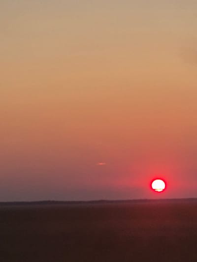 The image shows a sunset over a flat, dark landscape. The sun, a bright red orb with a distinct white halo at its bottom edge, is low on the horizon, casting a warm, gradient glow across the sky, transitioning from a pale peach at the top to a deep magenta near the horizon. A faint horizontal streak of light is visible in the sky above and to the left of the sun. The foreground is a dark, indistinct expanse, possibly indicating a field or prairie, with a subtle darker line suggesting a distant treeline or elevation. The scene appears to be located in an open, rural environment.