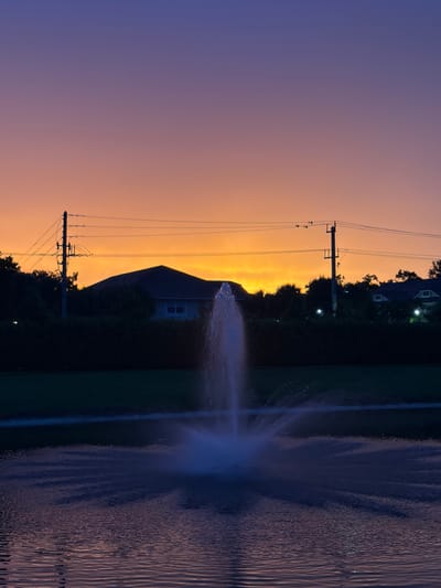 This vertical, low-angle, outdoor, full shot captures a dusk scene with a vibrant gradient sky transitioning from deep purple at the top to bright orange at the horizon. In the foreground, a body of water reflects the sky's colors, with a fountain erupting in the center, its white spray illuminated by unseen light sources. The fountain's spray is the primary subject of the foreground. Behind the water, a dark, dense line of foliage separates the water from the background. Above the foliage, a residential area is visible, silhouetted against the bright sky. A peaked-roof house with a light-colored facade and two windows is partially obscured by trees. Electrical poles and wires are strung across the middle ground, crossing in front of the house and trees. Several houses with illuminated windows are scattered throughout the background. The scene is set in Lauderdale Lakes, United States of America, likely in a suburban residential area with a common amenity like a community pond with a fountain. No people are visible. The lighting suggests twilight or just after sunset.