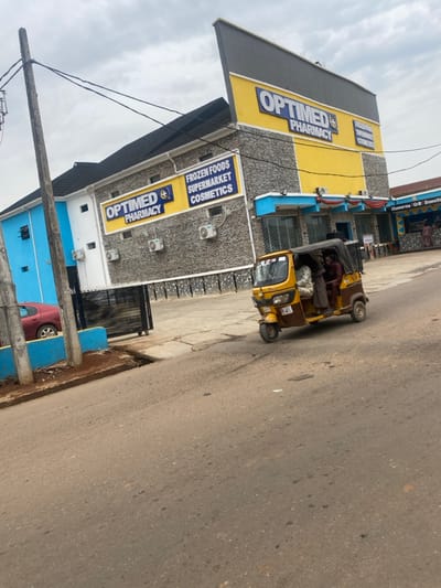 The scene depicts a daylight street view in the city of AlakIa, Nigeria, under an overcast sky. Dominating the right side is a two-story commercial building with a distinct yellow and gray facade. The upper section is yellow, displaying large blue lettering for "OPTIMED PHARMACY." The lower section features a gray textured or stone-like finish. Signs on the building also advertise "FROZEN FOODS," "SUPERMARKET," and "COSMETICS." Several exterior air conditioning units are mounted on the lower facade. Adjacent to the pharmacy, a smaller building with blue trim shows signage for "Shawarma Grill Smoothie," with indistinct figures of people visible in its vicinity.
In the midground, a brown and yellow three-wheeled auto-rickshaw (Keke Napep) is in motion, positioned on a cracked asphalt road, turning right. Two individuals are inside the auto-rickshaw; the driver, wearing a maroon-colored garment, is visible in the front, and a passenger, partly obscured, is seated behind. To the left, a tall wooden utility pole with multiple wires extends upwards. A red sedan is partially visible, parked behind a low blue barrier or wall on the left side of the frame. The road surface exhibits cracks and scattered debris.