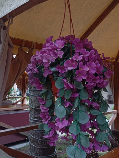 A hanging artificial flower arrangement with pink blossoms and green leaves is suspended from a wooden structure covered with a tan fabric awning. The arrangement is draped around a tree trunk wrapped with wire. In the background, there is a wooden railing, and furniture is visible. A timestamp "26.07.2026 15:40" is present in the lower right corner. The scene is located in the city of Verkhnyaya Balkariya, Russia.