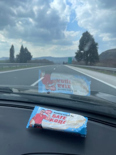 The image presents a view from inside a vehicle, looking forward through its windshield onto a multi-lane highway. The scene is set on a highway in a rural or semi-rural area, consistent with the region around Kipra, Bulgaria.
In the immediate foreground, a dark-colored vehicle dashboard is visible. Resting on this dashboard is a single, packaged wafer bar. The package features a light blue and white design with a cartoon illustration of a muscular man in a red polo shirt and sunglasses, flexing his bicep. Prominent Cyrillic text on the package reads "БАТЕ КОЦЕ" (Bate Kotse), with smaller text indicating "Всички обичат" (Everybody loves) and "Бялата вафла на" (The white wafer of). An inverted reflection of an identical or similar wafer bar package is visible on the vehicle's windshield, displaying fragmented Cyrillic text, including "КОЦЕ РУЛЕ" (Kotse Rule).
Beyond the vehicle's interior, a paved highway with white lane markings extends into the distance, suggesting at least two lanes of travel in the visible direction. A metal guardrail parallels the right side of the highway. In the midground, several trees are visible, including a prominent, tall dark green coniferous tree to the right of the road. A green road sign is faintly discernible further down the highway on the right. The background consists of rolling hills or mountains under a partly cloudy sky, showing patches of blue amidst a predominant grey cloud cover. The overall environment suggests an open road during daylight hours.