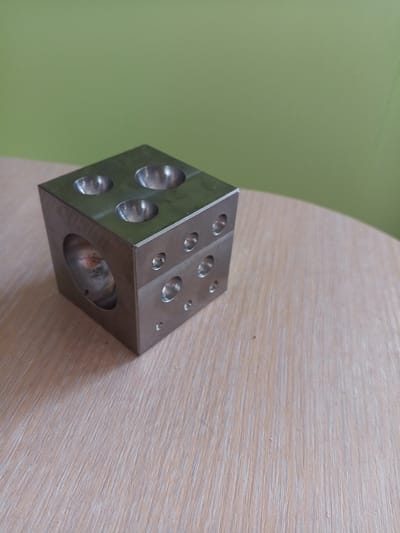 A metal cube with various indentations and holes sits on a light-colored wooden surface. The cube is positioned in the lower left quadrant of the image. The background is a solid, light green color. The image is taken from a slightly elevated angle looking down. The scene appears to be indoors, likely on a table. There is no indication of the city of Vilyuysk, Russia in the image.
