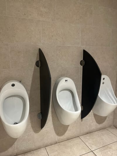 The image presents a modern public restroom interior, specifically a wall-mounted urinal arrangement, located in Dorno, Italy. Three sleek, white, teardrop-shaped urinals are mounted vertically on a light-colored tiled wall. Each urinal features a small, metallic flush mechanism or sensor with two circular elements.
Between the urinals are two dark, semi-circular shelves, also mounted on the wall. These shelves appear to be made of a dark, possibly wooden, material and are supported by two visible metallic brackets each. The wall itself is covered in rectangular, light beige tiles with a subtle, textured finish, laid in a horizontal pattern. Similar tiles are visible on the floor, suggesting a uniform aesthetic.
The scene is illuminated by an unseen light source, casting gentle shadows from the urinals and shelves onto the wall, emphasizing their form and depth. No people are present, and no activity is occurring; the urinals are empty and appear clean and well-maintained. The contemporary design of the fixtures and the clean tiling indicate a modern and functional public facility. There is no visible text in the image.