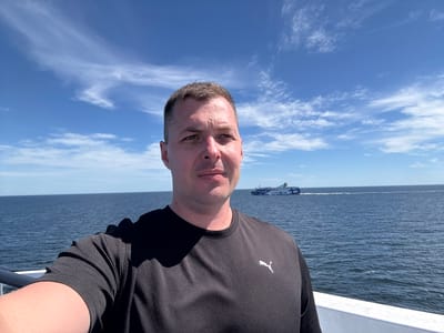 A man in a black Puma t-shirt takes a selfie on a ferry with the sea and sky in the background. He is looking towards the camera with a neutral expression. In the distance, a blue and white ferry can be seen sailing on the water under a cloudy blue sky. The sea is a deep blue, and the sky is a bright blue with wispy white clouds. The man is standing on the deck of a ferry, as indicated by the railing in the foreground.