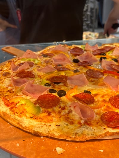 A close-up, overhead shot showcases a large, circular pizza topped with melted cheese, pepperoni slices, ham pieces, black olives, and sliced bell peppers in green, yellow, and red. The pizza rests on a wooden pizza peel, which is on an orange surface that appears to be a countertop or serving area. In the blurred background, out-of-focus individuals are visible, suggesting a public setting like a restaurant or food establishment. The overall lighting is bright, highlighting the textures and colors of the pizza. No specific details are available regarding the location in Phu Thuy, Vietnam, as this information is not discernible from the image itself.