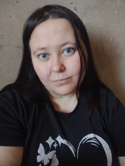 The image shows a close-up of a young woman with long, dark hair and blue eyes. She is wearing a black t-shirt with a white graphic design that appears to include a butterfly and stars. The background is a textured, neutral-colored wall. The image was taken in Sukhonogovo, Russia.