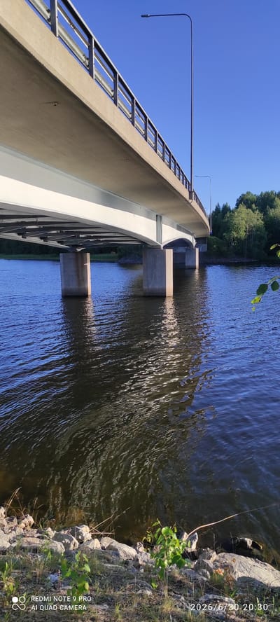 The image shows a bridge over a body of water. The bridge has a concrete walkway with a metal railing. A street light is visible on the left side of the bridge. The water is dark and appears to be a lake or river, with ripples on its surface. Trees and vegetation line the far bank of the water. The sky is clear and blue, indicating good weather. The scene is lit by bright sunlight, suggesting it is daytime. The overall impression is one of a peaceful, natural setting. The time of day is indicated as 20:30 on 2026/6/30. The text "REDMI NOTE 9 PRO AI QUAD CAMERA" is visible in the lower right corner.