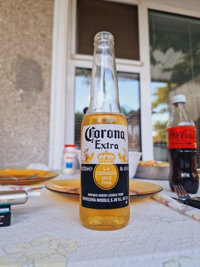 This media file is a close-up, slightly angled shot of a table setting outdoors, likely in a backyard or patio area. The primary subject is a bottle of Corona Extra beer, lying on its side on a patterned tablecloth. The beer bottle is mostly empty, with a small amount of liquid visible. The label is clearly legible, showing the "Corona Extra" logo, "LA CERVEZA MAS FINA," and "Alc. 4.5% vol." Text also indicates it is "BREWED UNDER LICENCE FROM CERVECERÍA MODELO, S. DE R.L. DE C.V."
In the background, a Coca-Cola bottle is visible, standing upright. A fork and knife are also present, suggesting a meal or drinks were being consumed. The setting appears to be daytime, with natural light illuminating the scene. Behind the table, a window frame and part of a building with a textured, light-colored wall are visible, indicating an outdoor or semi-outdoor location. The overall impression is one of a casual outdoor gathering. There are no people visible in the image. The location context provided is Ovoshtnik, Bulgaria.