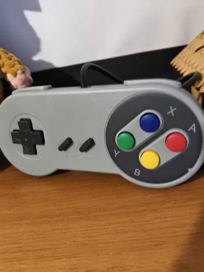 A close-up, eye-level, medium shot captures a gray video game controller resting on a wooden surface. The controller is positioned horizontally, with its face angled slightly upwards and towards the right. The directional pad, a black cross shape, is on the left side of the controller. To the right of the directional pad are two black buttons labeled "SELECT" and "START". The right side of the controller features four colored face buttons arranged in a diamond pattern: a green "Y" button, a blue "X" button, a red "A" button, and a yellow "B" button. The background is out of focus, showing a white surface above a black surface. The wooden surface appears to be a table or shelf. The scene is indoors, likely in a residential setting, and there are no visible individuals or animals. The lighting is even, suggesting an ambient light source. The image does not contain any information that would allow for identification of its location within the city of Mokshan, Russia.