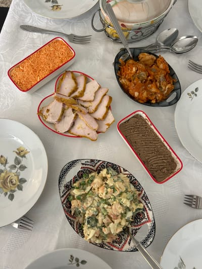 The image shows a table set for a meal. It appears to be a festive or special occasion setting, with multiple dishes arranged for sharing. The dishes include:

*   A dark-colored stew or braised dish in a black patterned serving bowl.
*   A rectangular dish of what appears to be pâté or a similar spreadable item, featuring a ridged surface. The dish has a white body with a red border.
*   A platter of sliced, cooked turkey or chicken breast. The platter has a white body and a red border.
*   A rectangular dish of grated carrots.
*   A salad with a creamy dressing containing what appears to be peas, carrots, and other vegetables, in an intricately patterned oval serving bowl.

The dishes are placed on a white tablecloth. There are also plates set around the table, each with a floral design. Silverware is visible next to the plates.

The location is not discernible from the image. There are no elements that identify this is Karmiel, Israel. The picture shows an indoor environment in what appears to be a dining room, focused entirely on the food setup.