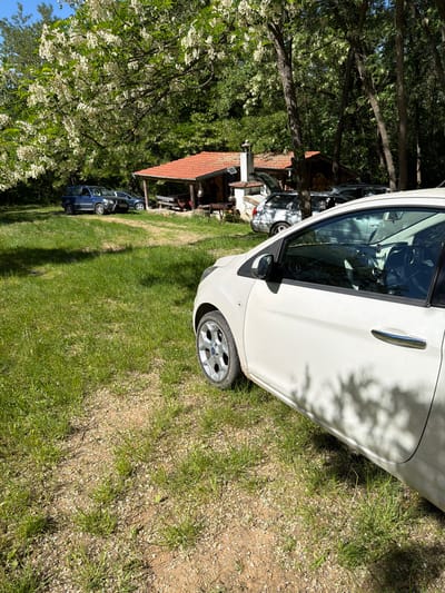 The image shows a sunny outdoor scene, possibly in a rural area of Kanchevo, Bulgaria. The foreground is dominated by a white car parked on grass. In the background, there is a rustic structure with a red-tiled roof, surrounded by trees and greenery. There is also another blue car parked further back. The setting suggests a residential or recreational area with elements of both natural and built environments. No people are visible in the photograph.