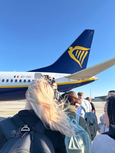 This image captures an outdoor scene at an airport, likely on the tarmac, bathed in bright daylight under a clear blue sky. A group of people are viewed from behind, appearing to queue or walk towards the rear entrance of a large passenger airplane, indicating a boarding process is underway.
The dominant object is a Ryanair aircraft, identifiable by its distinct dark blue tail fin featuring the airline's yellow harp logo. The plane's fuselage is white with a yellow stripe and blue livery. Clearly visible on the side of the fuselage is the aircraft registration "EI-IGI" positioned next to a small Irish flag, confirming its Irish registration. The people are moving towards an open door at the back of the aircraft.
In the foreground, closest to the camera, is the back of a person with long, blonde or light brown hair, wearing a dark top and a black backpack. A decorative hair clip, possibly made of white beads or pearls, is attached to a strap of their backpack. Further into the frame, other individuals with varied hair colors (including dark, brown, and red-brown) are seen, also from behind. They are dressed in casual clothing, such as sweatshirts and jackets, and some are carrying backpacks, including a prominent light green/teal one.
The bright sunlight suggests it is daytime, with clear weather conditions. The scene depicts passengers boarding a flight, likely a low-cost carrier, at an airport. Given the provided context, the event is taking place in Weeze, Germany.