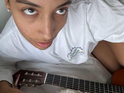 The image captures a close-up, high-angle view of an individual positioned indoors. The person has dark hair and brown eyes, looking directly upwards towards the camera. They are wearing a white, long-sleeved t-shirt. On the upper right chest area of the t-shirt, a graphic logo is visible, featuring text that includes "CLEANERS Uncle Paulie's Sandwich Muffuletta Sandwich Los Angeles August 21, 2022". A small, gold-colored, heart-shaped stud earring is present in the person's left earlobe. Below the individual's torso, the fretboard and headstock of a classical guitar are prominent. A silver-colored capo is clamped onto the guitar's headstock, over the strings. The immediate environment appears to be a light-colored, soft surface, consistent with bedding or fabric. The specific location in Maisons-Alfort, France, cannot be determined from the visual information present in the image.