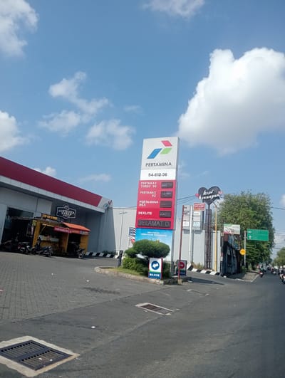 The image depicts a bright, sunny day with a clear blue sky filled with scattered white clouds. In the foreground, a gray asphalt road leads into a gas station. To the left, a Pertamina gas station with a red and white awning is visible, housing a food stall selling kebab. Several motorcycles are parked in front of the stall.

Dominating the center of the image is a tall Pertamina gas price sign displaying current fuel prices. Adjacent to it, a sign for "kopi kenangan" (memory coffee) is illuminated. Further down the road, a green traffic sign indicating directions can be seen. The scene appears to be located in the city of Sidoarjo, East Java, as suggested by the Pertamina brand, a prominent Indonesian state-owned oil and gas company. The environment is a typical urban street scene, with traffic visible in the distance, suggesting a busy day.