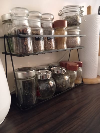 In the city of Baden, Austria, a wooden countertop displays a two-tiered black metal spice rack filled with various spices in glass jars. The top shelf holds five jars. From left to right, the first jar contains green herbs, the second contains small brown nuts, the third contains a spice with small brown seeds, the fourth is labeled "kori" and contains seeds, and the fifth contains a yellowish spice. Behind these, a larger glass jar with a clamp lid contains dried herbs, and a roll of white paper towels is positioned to the right. The bottom shelf holds six jars. The leftmost jar is large with a silver lid and contains black peppercorns. The next three jars contain spices and are topped with reddish-brown lids. The remaining two jars on the bottom shelf are partially visible. A white appliance, possibly a coffee maker, is situated to the left of the spice rack. The background is a light-colored wall.