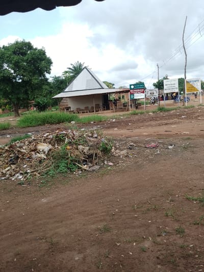 This image depicts an outdoor scene in Adikpo, Nigeria, likely during the daytime. A large pile of garbage is prominently featured in the foreground, consisting of various types of refuse, including plastic bags, paper, and dried organic matter. Beyond the garbage, a dirt road leads to a cluster of buildings.
One of the structures appears to be a small market or roadside stall with a corrugated metal roof supported by wooden posts. Several people are seated under this shelter. In the background, there are more buildings, including what seems to be a school, indicated by signage that reads "RCM PRIMARY SCHOOL."
Several other signs are visible, including one that says "NKST CHURCH HILAKAAN" and another that reads "GOVERNMENT OF BENUE STATE MINISTRY OF EDUCATION AREA EDUCATION OFFICE." These signs suggest a community setting with religious and educational institutions.
The overall impression is one of a rural or semi-urban environment. The weather appears to be overcast, with a cloudy sky. The presence of a large pile of unmanaged waste suggests potential environmental challenges in the area.