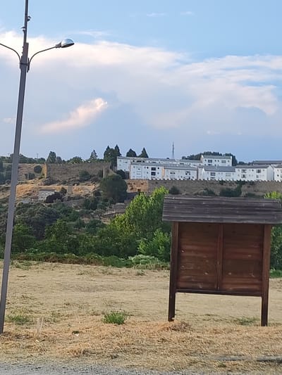 The image depicts a scene in the city of Béjar, Spain, likely taken during daylight hours. In the foreground, a wooden information board stands on two legs, its surface weathered and brown. Behind it, a dry, grassy field extends towards a hillside. The hillside is covered in vegetation and features several buildings, including what appears to be an old stone wall or fortification. Above the wall, a row of white buildings with multiple windows is visible. Further in the distance, a tall, slender tower or antenna can be seen against a sky with scattered clouds. To the left of the frame, a tall, grey lamppost with a curved arm supporting a light fixture is present. The overall impression is of an outdoor public space, possibly near historical ruins or a viewpoint.