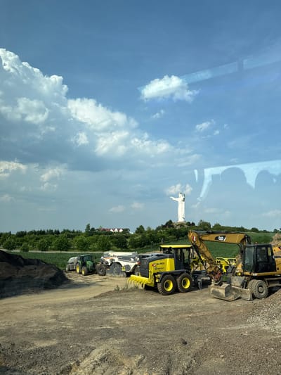 In Osogóra, Poland, an outdoor scene features a construction site with various heavy machinery, including a CAT excavator, a yellow compactor, and a green tractor. The ground is covered with dirt and gravel. In the background, a large white statue of Jesus Christ stands on a hill, with a few houses visible behind it amidst lush green trees. The sky above is a bright blue with scattered white clouds. Reflections of what appear to be buildings or structures are visible on a glassy surface in the upper portion of the image.