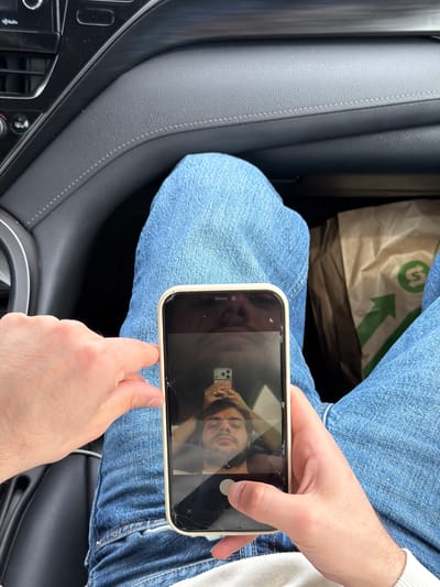 A person sits in the passenger seat of a car. The person is wearing blue jeans. The person holds a damaged white smartphone displaying a selfie photo. A brown paper bag with green lettering rests on the person's lap. The car has a grey interior. The location is Tserovani, Georgia, based on the bag's logo.