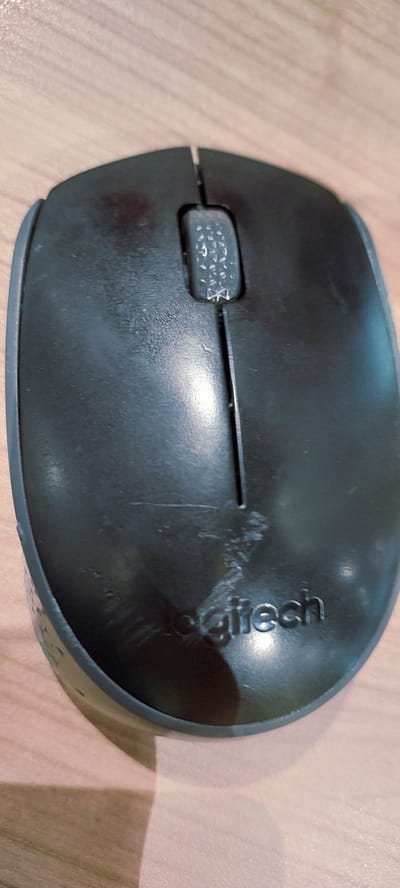 The media file shows a close-up, overhead shot of a black Logitech wireless mouse resting on a wooden surface. The mouse is positioned diagonally from the bottom left to the top right of the frame. The scroll wheel is visible on the left side of the mouse, with a textured pattern. The "Logitech" logo is embossed on the right side of the mouse, though it appears somewhat worn or obscured by smudges. The wooden surface beneath the mouse has a light brown, grained texture. The lighting suggests an indoor setting, possibly with overhead artificial light creating a shadow on the right side of the image, extending towards the bottom right. There are some scuff marks and smudges visible on the surface of the mouse, indicating it is a used item. No people are visible in the image. The scene is a simple still life of an electronic device.