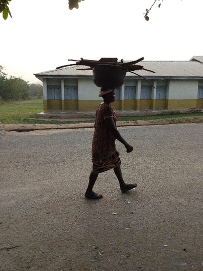 An adult female is depicted walking leftwards on an asphalt road in Igbo Ijaiye, Nigeria. She wears a patterned dress and dark sandals. A large, dark basin or pot, filled with cut firewood, is balanced on her head, with a light-colored cloth providing support beneath it. In the background, a single-story building features a light-colored upper facade and a darker, possibly yellow, lower section, topped by a light gray corrugated metal roof. The building's facade contains several rectangular, dark blue-gray door or window closures. To the left of the building, a field of green grass with sparse trees is visible. The sky is uniformly bright and hazy. Out-of-focus tree branches are present in the upper left corner of the frame.