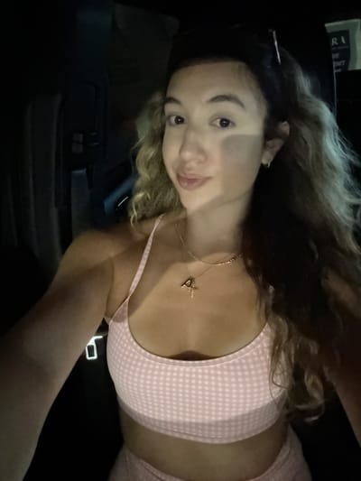 The image shows a young woman lying down and looking at the camera. She has long, wavy, blonde and brown hair. She is wearing a light pink and white checkered bikini top and matching shorts. Around her neck, she has a delicate gold necklace with a small charm. The scene appears to be inside a vehicle, with a dark interior and a black car seat visible. The lighting suggests it is nighttime. There is some visible text on a white object in the upper left corner of the image that appears to say "RAI" or similar, but it is largely obscured. The woman's expression is a slight smile, with her head tilted. The overall mood of the image is casual and relaxed.
