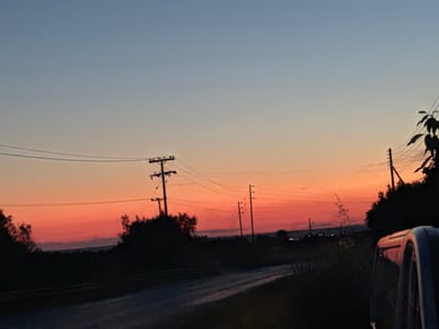 A dusk scene shows a road through the city of Γερακινή, Greece. Silhouetted vegetation flanks the road, with telephone poles and wires stretching across the sky. The sky is gradated from blue at the top to orange and red near the horizon. The road appears to be paved. A vehicle's side mirror is visible in the lower right. Distant lights appear on the horizon.