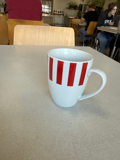 A white mug with red stripes sits on a light-colored, speckled surface, possibly a table, in a cafeteria or dining hall. The mug is positioned to the right of the frame, with its handle visible on the right side. In the background, several wooden chairs are partially visible, along with other people seated at tables. One person is wearing a red shirt, another is wearing a black t-shirt, and a third is wearing a grey hoodie and glasses. The environment appears to be indoors, with artificial lighting. The overall scene suggests a casual dining or study setting. The image was captured in Ganderkesee, Germany.