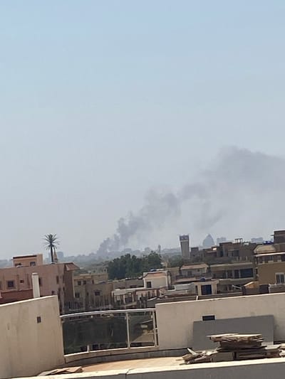 A view from a rooftop in Omdurman, Sudan, showing a dense cloud of smoke rising in the distance. The smoke is visible against a pale blue sky. The rooftop is partially obscured by a tall building in the foreground, and there are other buildings in the distance, including a tall minaret. There is a single palm tree visible near the base of the tall building in the foreground.