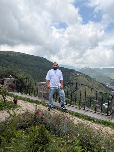 A man stands on a paved walkway, leaning against a low stone wall with a black metal railing. He wears a white t-shirt with a black logo and light blue jeans. Behind him, a lush green mountainside stretches into the distance, dotted with trees and scattered buildings. The sky is overcast with large, white clouds. In the foreground, there are rose bushes with red and pink flowers, and various other plants. The location is the viewpoint overlooking the city of Goris, Armenia, known for its dramatic landscape of green hills and valleys.