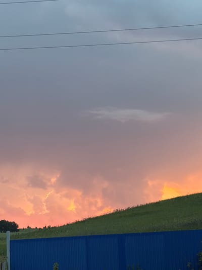 The image depicts a rural landscape at dusk, likely in or near the village of Subkhankulovo, Russia. The sky is dominated by a dramatic sunset with hues of pink, orange, and purple in the clouds. Three parallel power lines stretch horizontally across the upper portion of the frame. Below the sky, a grassy hill slopes upwards from left to right. On the far left of the hill, a dark tree stands. A vibrant blue fence runs horizontally across the bottom of the image, partially obscuring the base of the hill. The scene is peaceful and appears to capture the natural beauty of the countryside during twilight.
