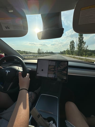The image provides a driver's perspective from within a moving Tesla vehicle on a highway near Kapitan Andreevo, Bulgaria, under bright daytime conditions. The driver's hands grip the steering wheel, with the right arm, adorned with a black braided bracelet, prominently visible. Tattoos are discernible on the driver's left leg, partially covered by light-colored shorts. A passenger's right knee is visible in the lower right foreground. Both driver and passenger sun visors are in the upright position, displaying multi-language warning labels. The central Tesla touchscreen displays a speed of "88", a navigation map indicating a route with 7.9 km remaining, an outside temperature of "14 C", and an Autopilot visualization showing the car within its lane with a detected vehicle ahead. A media player interface is partially visible at the bottom of the screen. The center console contains an open storage compartment with crumpled paper or tissues. Through the windshield, a multi-lane asphalt highway stretches into the distance, flanked by green hills, trees, and guardrails. Tall, slender trees are visible along the right side of the road. A bridge or overpass is discernable on the distant horizon. The sky is blue with scattered white clouds, and the low sun is directly ahead, casting bright glare and lens flares across the windshield.