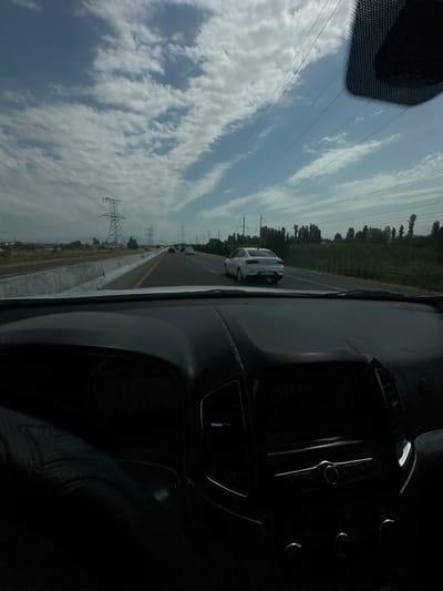 The image depicts a view from inside a vehicle traveling on a highway. The perspective is from the driver's seat, showing the dashboard and the windshield. Outside the vehicle, the highway stretches ahead, with other cars visible in the distance. Large power line towers and transmission lines are present along the side of the road, indicating a rural or semi-urban area. The sky is partly cloudy, with white clouds scattered across a blue sky. Trees and vegetation line the side of the highway in the background. The interior of the car appears dark, with the dashboard console and a portion of the steering wheel visible. The overall scene suggests a daytime journey on a highway, likely in a location with significant electrical infrastructure. The provided context specifies the location as the city of Istiqlol, Uzbekistan.