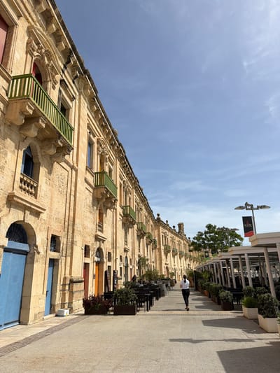 This photograph captures a street scene in Floriana, Malta, showing a row of historic buildings. The buildings, constructed from light-colored stone, feature ornate architectural details such as decorative carvings and distinct window frames, some with green shutters or bars.

A sidewalk runs along the side of the buildings, with chairs and tables from a restaurant, potted plants and a passerby walking away from the camera, appearing to be the only person in the scene. A street lamp with a black and red banner is also visible. The sky is predominantly blue with some wispy clouds.